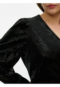 Robe noire avec un col en V et de longues manches bouffantes, confectionnée dans un tissu texturé avec un motif perforé réfléchissant, augmentant l'intérêt visuel.