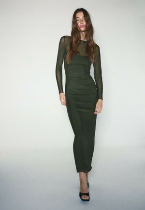 Femme aux longs cheveux bruns portant une robe vert foncé ajustée avec des manches longues transparentes et des talons noirs ouverts à l’avant, debout devant un fond clair uni.