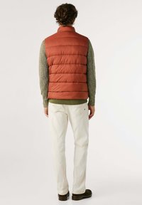 Gilet imbottito in arancione ruggine con texture a righe orizzontali, abbinato a un maglione in maglia verde e pantaloni crema. Collo fissato, senza hardware visibile.