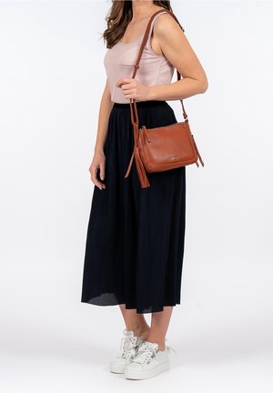 Kobieta w jasnoróżowym topie na ramiączkach i czarnej plisowanej spódnicy, trzymająca brązową skórzaną torebkę na ramię z detalem w postaci chwosta.