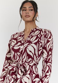 Langärmliges Kleid mit einem bordeaux- und cremefarbenen Blätterdesign, einer Knopfleiste auf der Vorderseite und einem gebundenen Taillenbereich für eine definierte Form. Glatte Stofftextur.