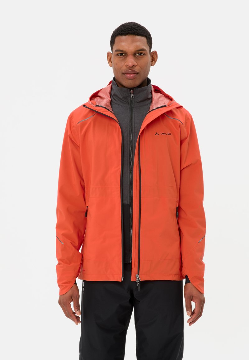 Man in een heldere oranje hoodie over een grijze zip-up top en zwarte broek, staand tegen een effen witte achtergrond.