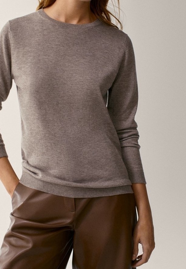 Femme portant un pull taupe à manches longues et un pantalon en cuir marron, debout avec une main dans la poche devant un fond neutre.