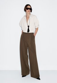 Persoon in een oversized wit shirt, met wijde bruine broek, zwarte puntige schoenen, een zonnebril en een lange zwarte tasselketting, staande tegen een witte muur.