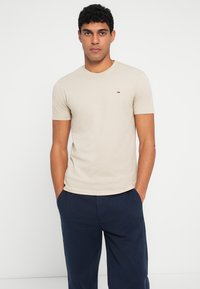 Μπεζ βαμβακερό t-shirt με κοντά μανίκια, στρογγυλή λαιμόκοψη και μικρό κεντητό λογότυπο στο στήθος, σε συνδυασμό με σκούρες μπλε παντελόνες.