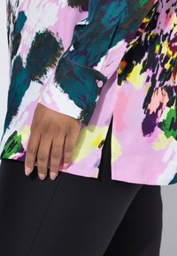 Multicolored long-sleeve blouse in pink, green, and yellow patterns, with a buttoned cuff and side slits, paired with black leggings.