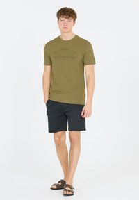 Vihreä oliivinvärinen puuvillapaita, jossa on painettu slogan, yhdistettynä mustiin shortseihin ja ruskeisiin sandaaleihin. Paita on yksinkertainen muotoilultaan ja rento istuvuudeltaan.