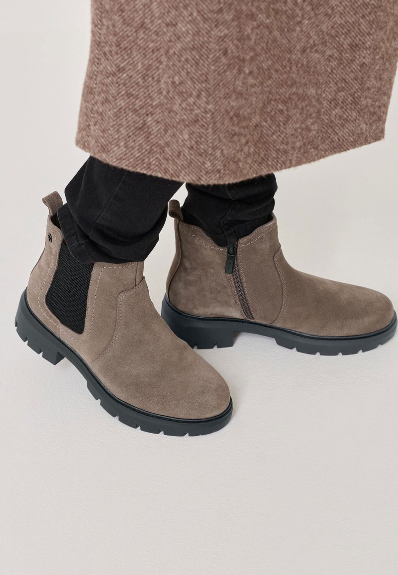 Bocanci scurți din suede maro, cu un panou elastic lateral negru și fermoar, având o talpă texturată pentru aderență și un design minimalist.