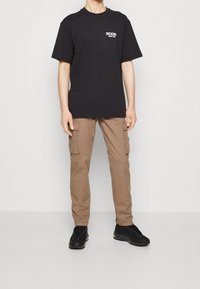 Schwarzes T-Shirt mit kurzen Ärmeln und weißem Druck, kombiniert mit beigen Cargo-Hosen mit Seitentaschen und schwarzen Sneakern mit strukturierten Sohlen.