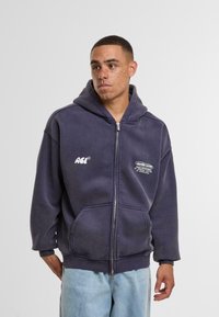 Sudadera con cremallera de color gris oscuro en tejido suave, con un bolsillo frontal, diseño oversize y texto impreso en blanco en el pecho y la manga.