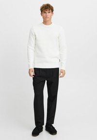 Pull en tricot texturé blanc avec un col rond, associé à un pantalon noir et des chaussures sombres, sur un fond clair uni.