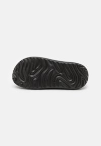 Semelle de chaussure en caoutchouc noir avec un motif de bande de roulement ondulé, présentant une texture lisse et un branding subtil sur le côté.