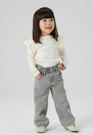 Maglietta bianca a maniche lunghe con coste e spalle arricciate, abbinata a pantaloni in denim grigi larghi e scarpe slip-on in tessuto chiaro.
