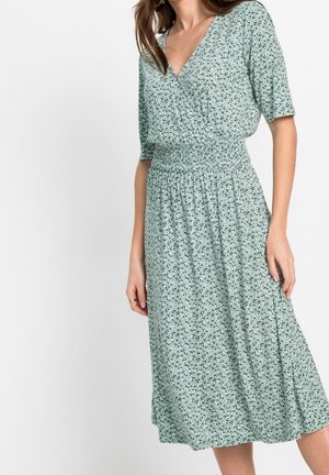 Donna che indossa un abito midi floreale verde chiaro con maniche corte e vita arricciata, in piedi su uno sfondo bianco uniforme.
