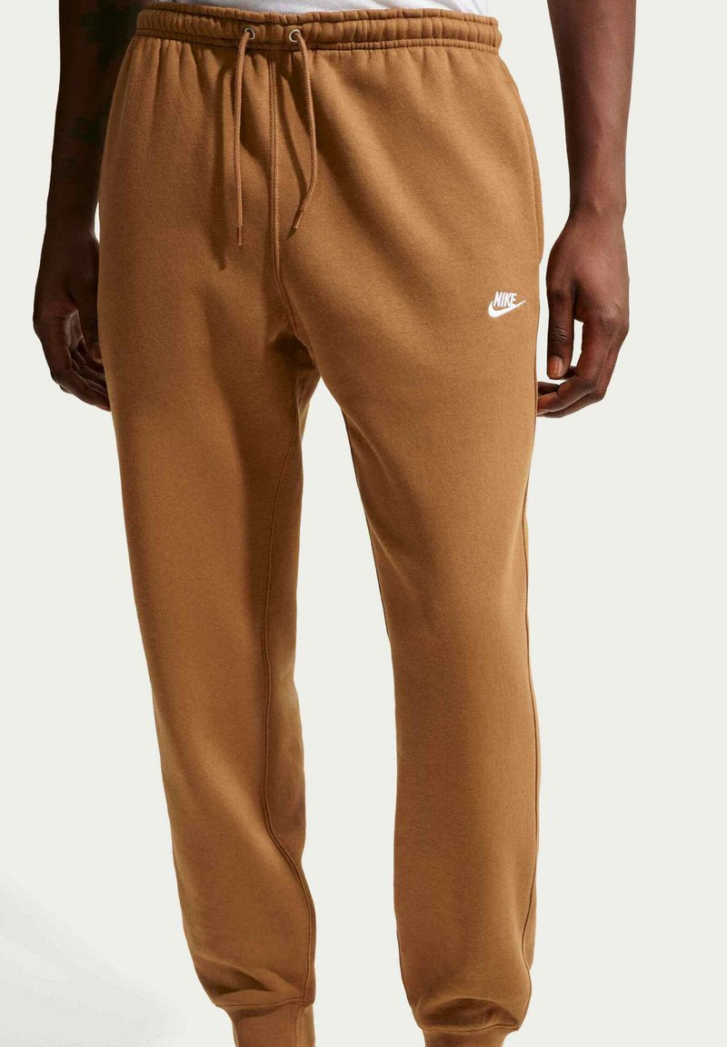 Pantalones deportivos marrones de Nike con cintura elástica, cordón y bolsillos laterales. Tela suave, piernas ajustadas y logo blanco destacado en el muslo.