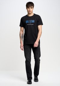 Camiseta de algodón negra con texto azul "BIG STAR" y eslogan "Tu Vida. Tus Jeans.", combinada con jeans negros y zapatillas negras.