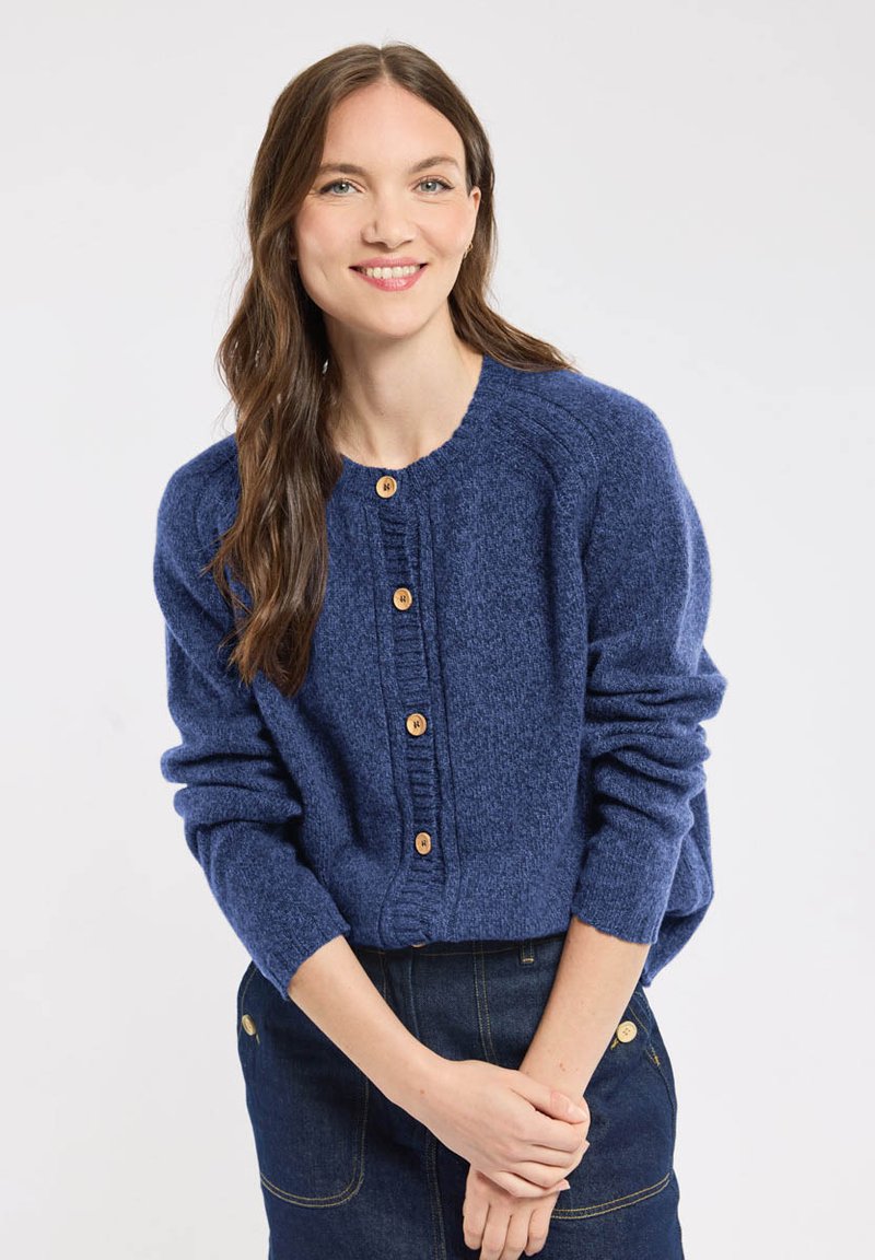 Gilet en tricot bleu avec un col rond, cinq boutons en bois et des poignets côtelés. Porté sur une jupe en denim noir avec des accents dorés.