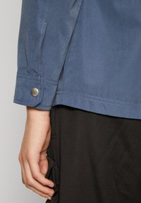 Blue fabric jacket close-up, featuring a snap button cuff, visible stitching, and a smooth texture. The hand is positioned on a dark garment.