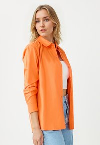 Camicia arancione con collo, maniche lunghe e vestibilità rilassata. Indossata sopra un crop top bianco, abbinata a jeans azzurri chiari.