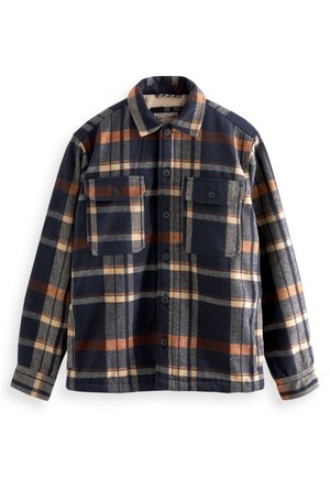 Mężczyzna noszący zapinaną na guziki koszulę kurtkę w kratę w kolorach granatowym, beżowym i brązowym z dwoma zapinanymi na guziki kieszeniami na piersiach.