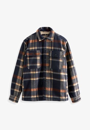 Uomo che indossa una giacca camicia a maniche lunghe a quadri abbottonata, nei colori navy, beige e marrone, con due tasche sul petto con bottoni.