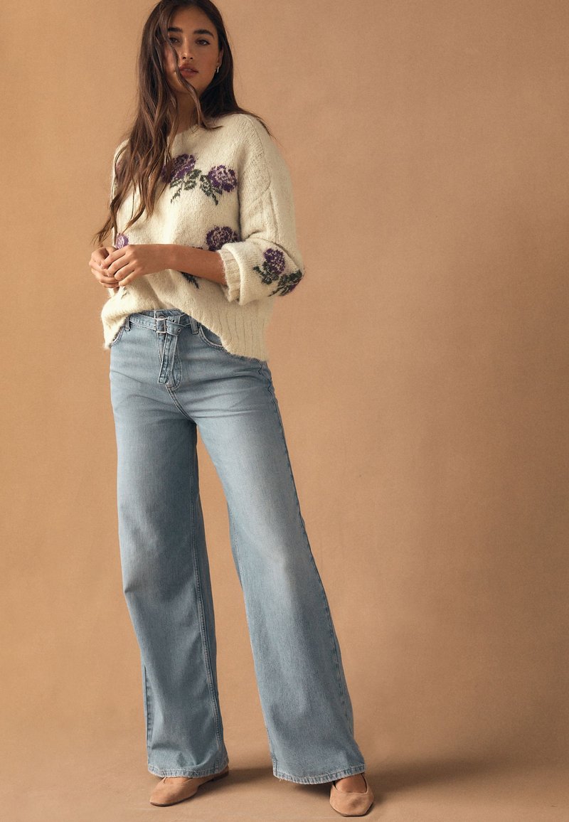 Femme debout portant un pull crème avec des fleurs violettes, un jean pantalon large bleu clair et des chaussures beige sur un fond beige.