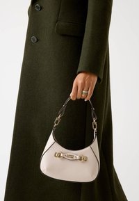 Beige Handtasche mit goldfarbenen Beschlägen und schwarzem Besatz, die eine gewölbte Silhouette aufweist und über einen abnehmbaren Tragriemen verfügt, gehalten von einer Hand in einem olivgrünen Mantel.