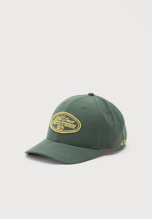 Mørkegrøn baseballkasket med gul broderet patch med teksten "Worldwide Sportswear Classic Varsity Aesthetic" på frontpanelet.
