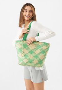 Die gewebte Tragetasche zeigt grüne und beige geometrische Muster, robuste grüne Träger und einen goldenen runden Anhänger.