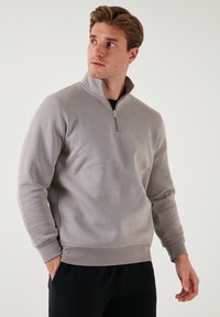 Sudadera gris con cremallera y alto cuello, hecha de una tela suave. Presenta una cremallera frontal y puños acanalados, ofreciendo un ajuste relajado.