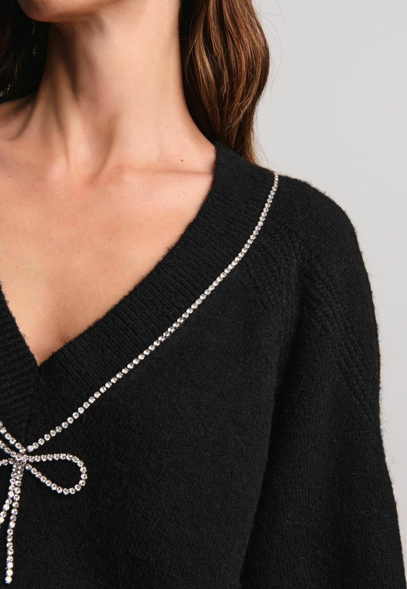 Femme portant un pull en maille noire avec un décolleté en V profond accentué par une bordure en nœud strass scintillant le long du bord.