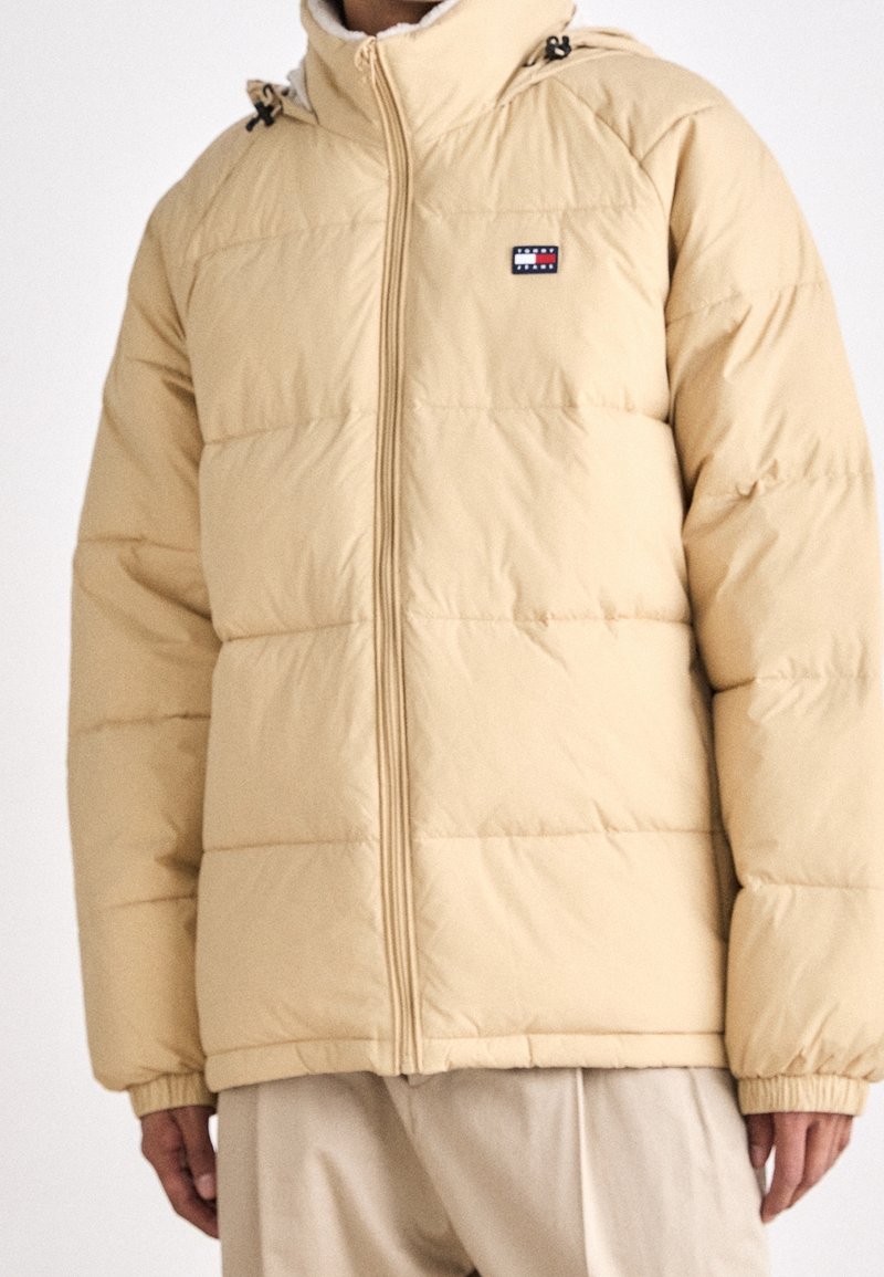 Doudoune beige avec fermeture éclair frontale et petite écusson rectangulaire sur le côté gauche de la poitrine, portée avec un pantalon beige assorti.