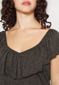 Top cinza escuro com decote em V e detalhes em babados. O tecido suave e texturizado destaca o design dos ombros e do decote.