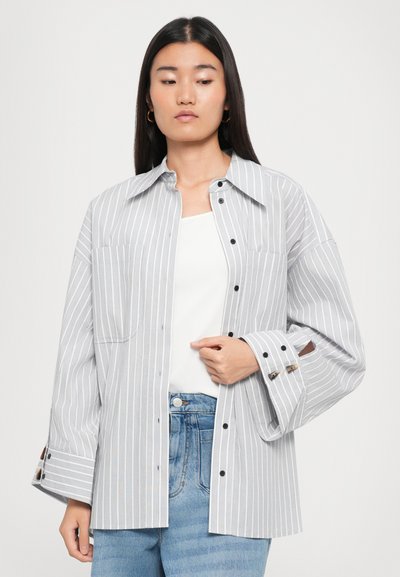 Gray and white striped button-up shirt with a relaxed fit, large collar, front pocket, and buttoned sleeve cuffs. Worn over a white top.