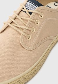 Bézs vászon sneaker, kerek orral, pamutszíjakkal és négy fém fűzőlyukkal. Jellemzője a juta borítású talp, amely textúrát és részletet ad.