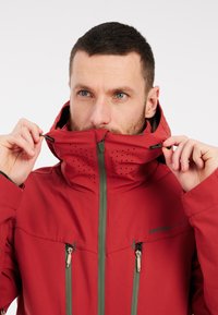 Rote wasserdichte Jacke mit hohem Kragen, verstellbare Kapuze, mit Reißverschlüssen versehenen Taschen und perforierten Details im oberen Bereich.