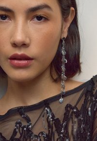 Silver dangle earrings with circular charms, exhibiting a delicate chain design. Model wears a black sequined top showcasing a sheer texture.