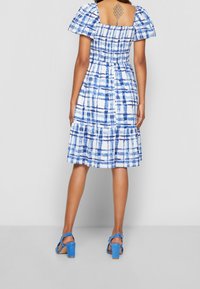 Blå och vit rutmönstrad klänning med puffärmar, smockad kropp, flerlagd fåll och öppen rygg. Bärs med blå högklackade sandaler.