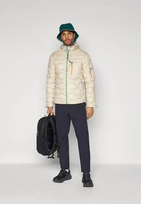Beige donsjack met doorgestikt patroon, groene rits, oranje accenten, gecombineerd met marineblauwe broek en zwarte rugzak, gedragen met een teal-colored bucket hat.
