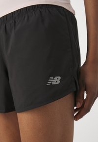 Pantalones cortos deportivos negros hechos de tela ligera, con una cintura elástica y un logo plateado de New Balance en el lado inferior.