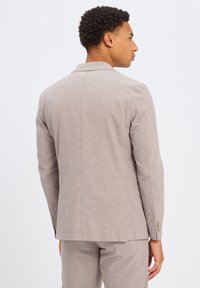 Lichtgrijze blazer met een getailleerde pasvorm, enkelvoudig knoopdesign, gladde textuur en subtiele naaiafwerkingen. Accentuerend met gouden knopen.