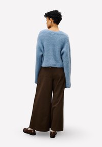 Frau steht mit dem Rücken zur Kamera, trägt einen hellblauen, flauschigen Pullover, weite braune Hosen und dunkle braune Slipper vor einem weißen Hintergrund.