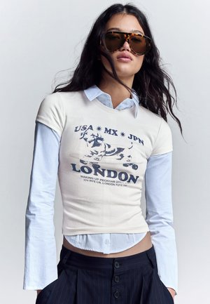 Femme portant de grandes lunettes de soleil écaille de tortue, chemise blanche superposée et t-shirt avec un chien sur un skateboard et le texte "USA MX JPN LONDON".