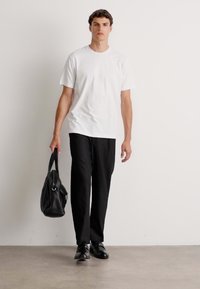 Un homme marche vers la caméra, portant un t-shirt blanc, un pantalon noir, des chaussures noires et portant un sac en cuir noir dans la main gauche.