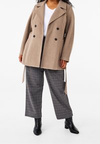 Syndig person i en beige dubbelknäppt kappa, vit skjorta, grå rutiga vida byxor och svarta ankelboots på en vit bakgrund.