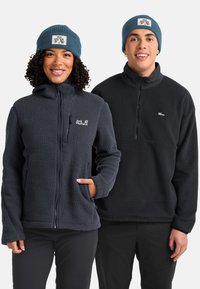 Glimlachende vrouw en man die blauwe mutsen en zwarte Jack Wolfskin fleece jassen dragen, naast elkaar staand tegen een witte achtergrond.