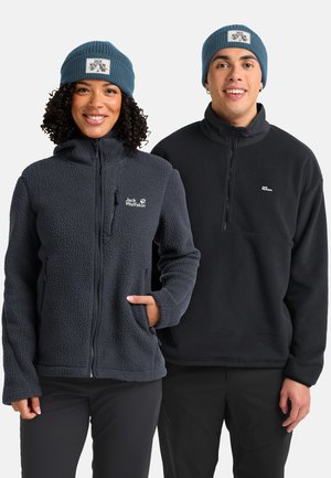 Smiling woman and man wearing blue beanies and black Jack Wolfskin fleece jackets, standing side by side against white background.