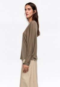 Dolgi rokavi, olivno zeleni top iz mehkega materiala, ki ima sproščen kroj z rahlo velikimi rokavi, v kombinaciji s bež krilom.