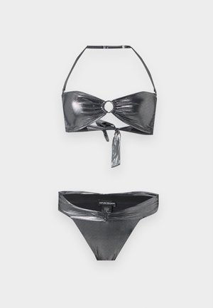 Jonge vrouw staat blootsvoets, draagt een metallic grijze tweedelige bikini tegen een effen witte achtergrond, handen losjes in elkaar gevouwen.