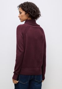 Burgunderfarbener Strickpullover mit hohem Kragen, strukturierten Schulterakzenten, gerippten Bündchen und Saum. Getragen mit blauen Jeans.